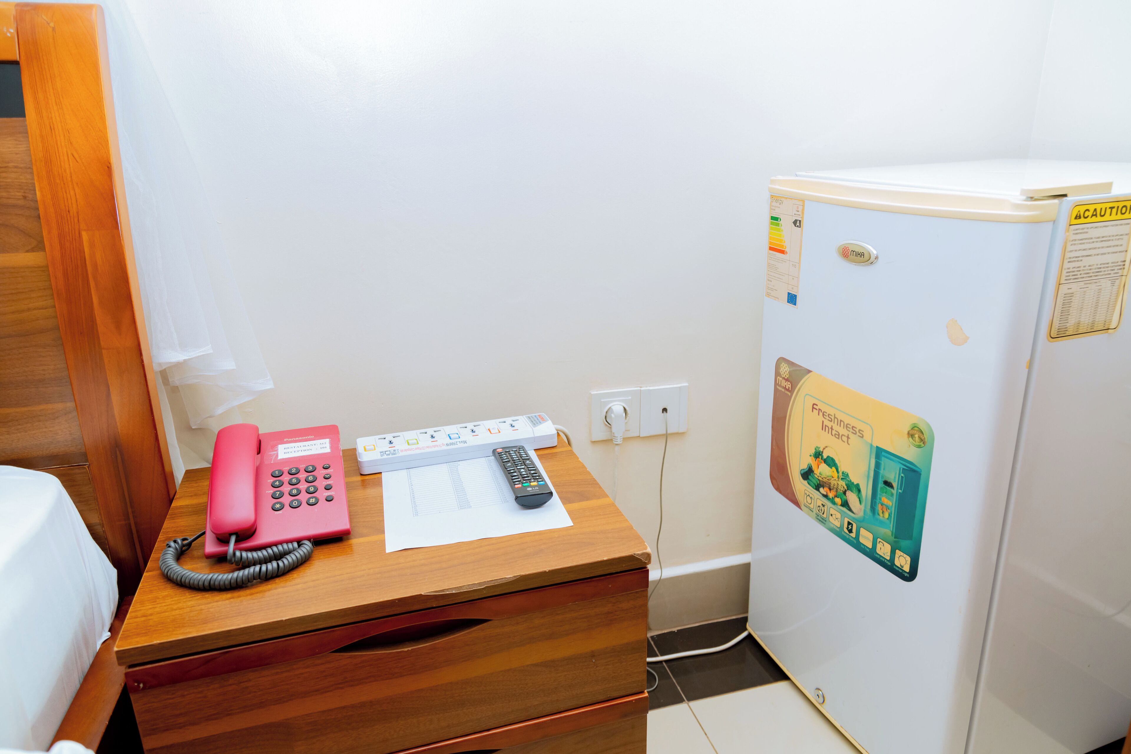 A wooden desk with a pink telephone, a remote control, and a notepad. A white refrigerator with a sticker is beside the desk.