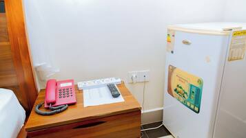A wooden desk with a pink telephone, a remote control, and a notepad. A white refrigerator with a sticker is beside the desk.