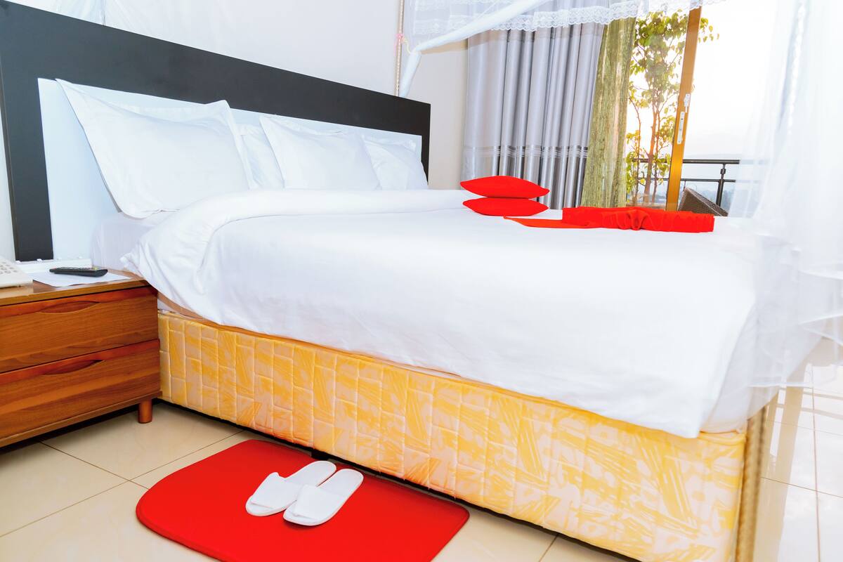 Una cama bien hecha con almohadas blancas y rojas, una mesita de noche de madera con un control remoto y una alfombra roja en el suelo.
