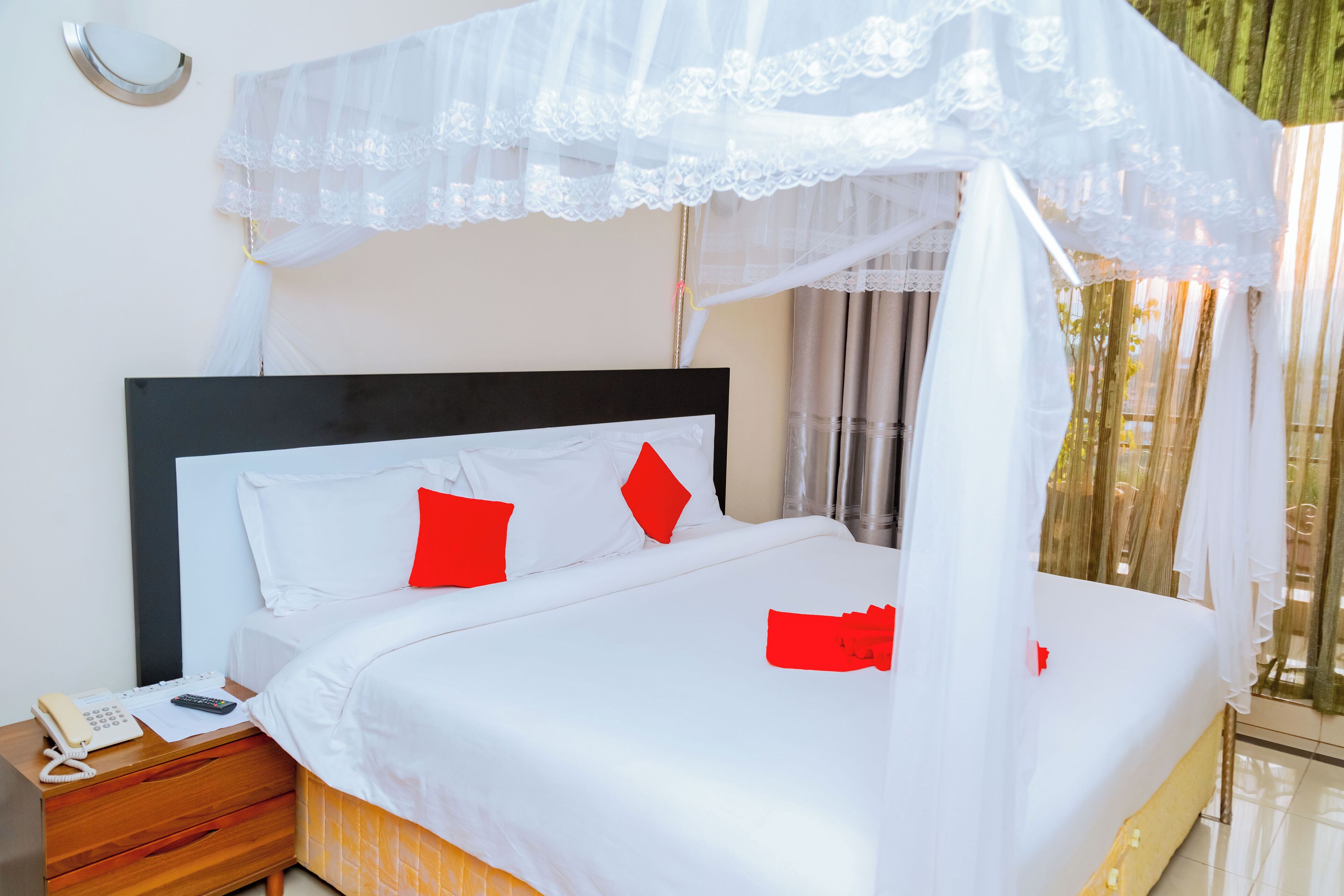 A neatly made bed with white bedding and red pillows, a bedside telephone, and a canopy with lace detailing.