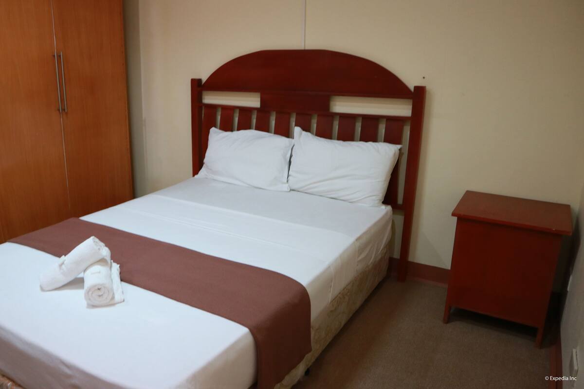 A neatly made bed with white linens and a brown bedspread, a wooden wardrobe, and a small wooden nightstand with a red top.