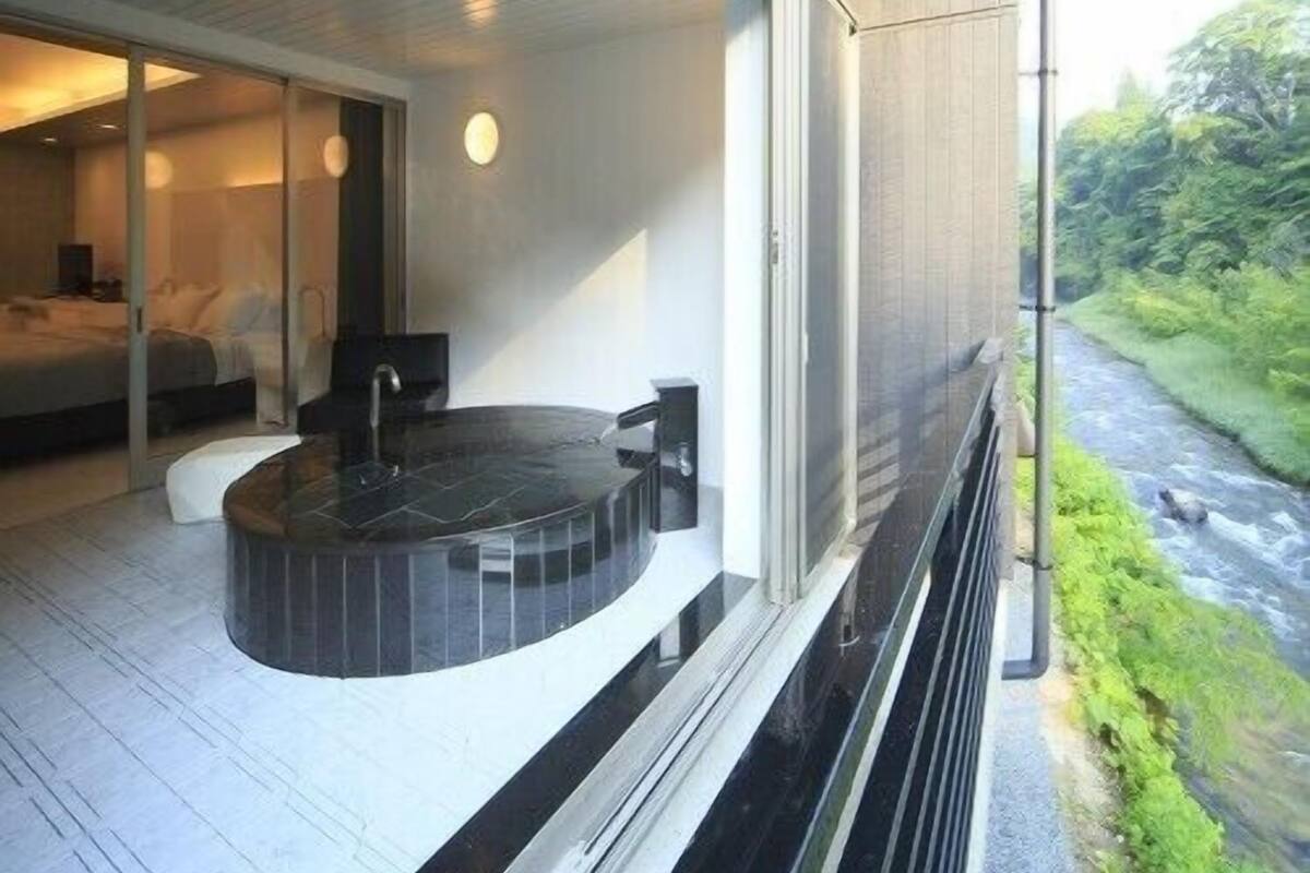 A modern hotel room with a circular hot tub, a large window overlooking a river, and a separate bedroom with a bed and a nightstand.