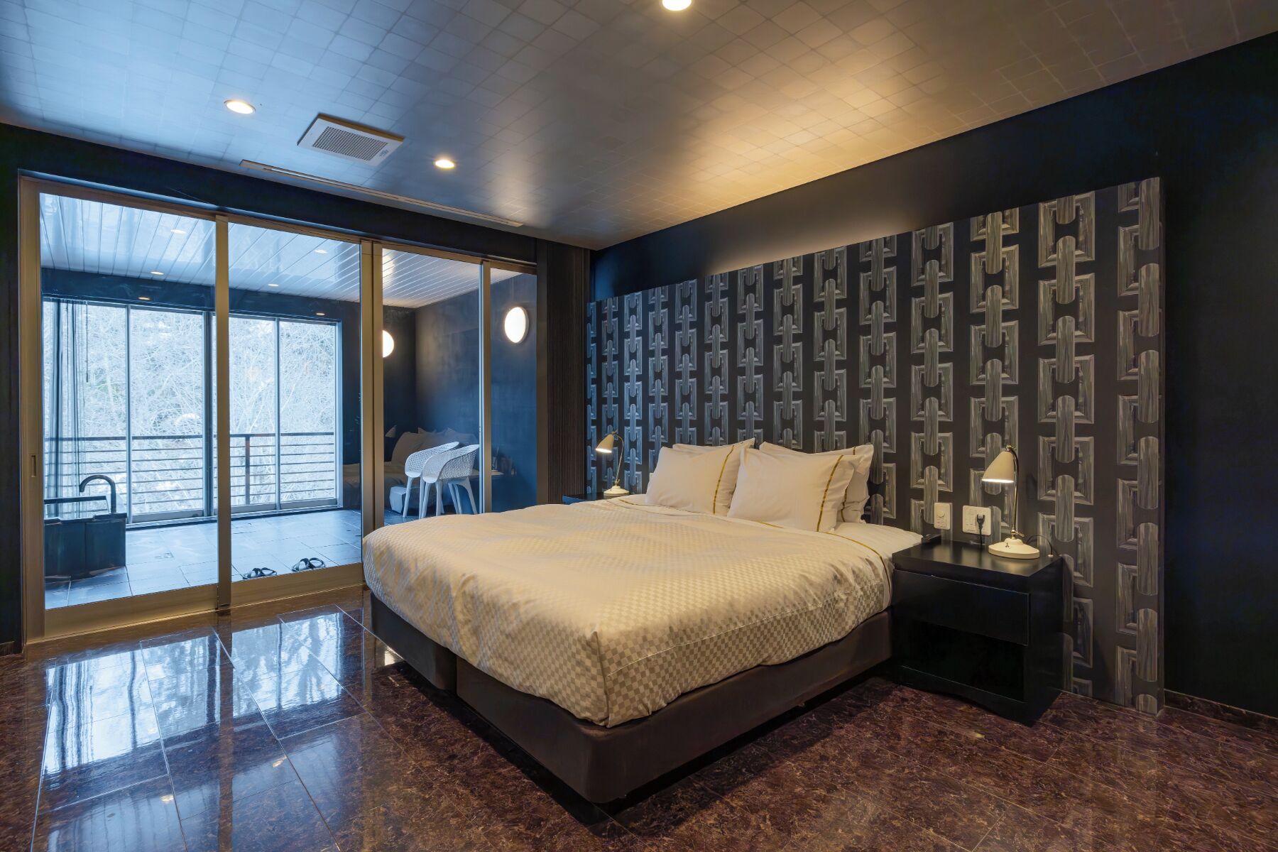 Une chambre moderne avec un grand lit, une table de chevet et une vue sur l’extérieur, accessible par une porte vitrée.