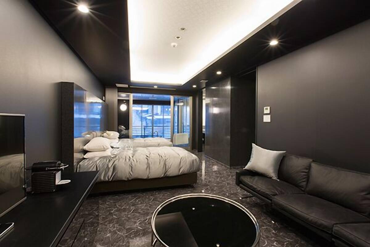 A modern bedroom with a large bed, a black leather sofa, and a glass-enclosed window.