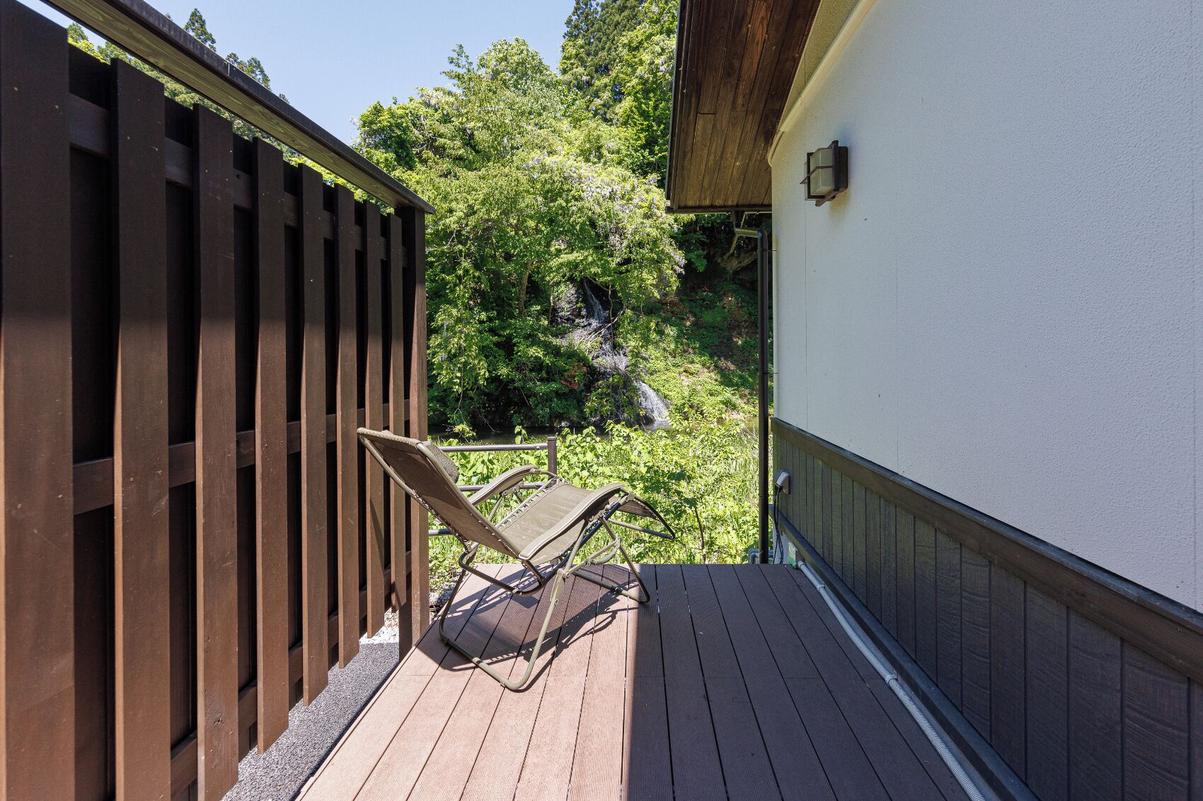 A wooden deck with two lounge chairs, a white building wall, and a wooden fence.