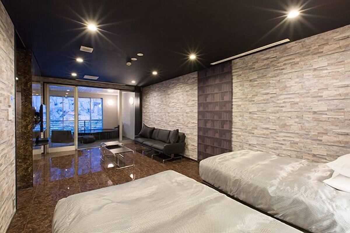 A modern hotel room with a stone-textured wall, a glass door leading to a balcony, a sofa, and two beds.