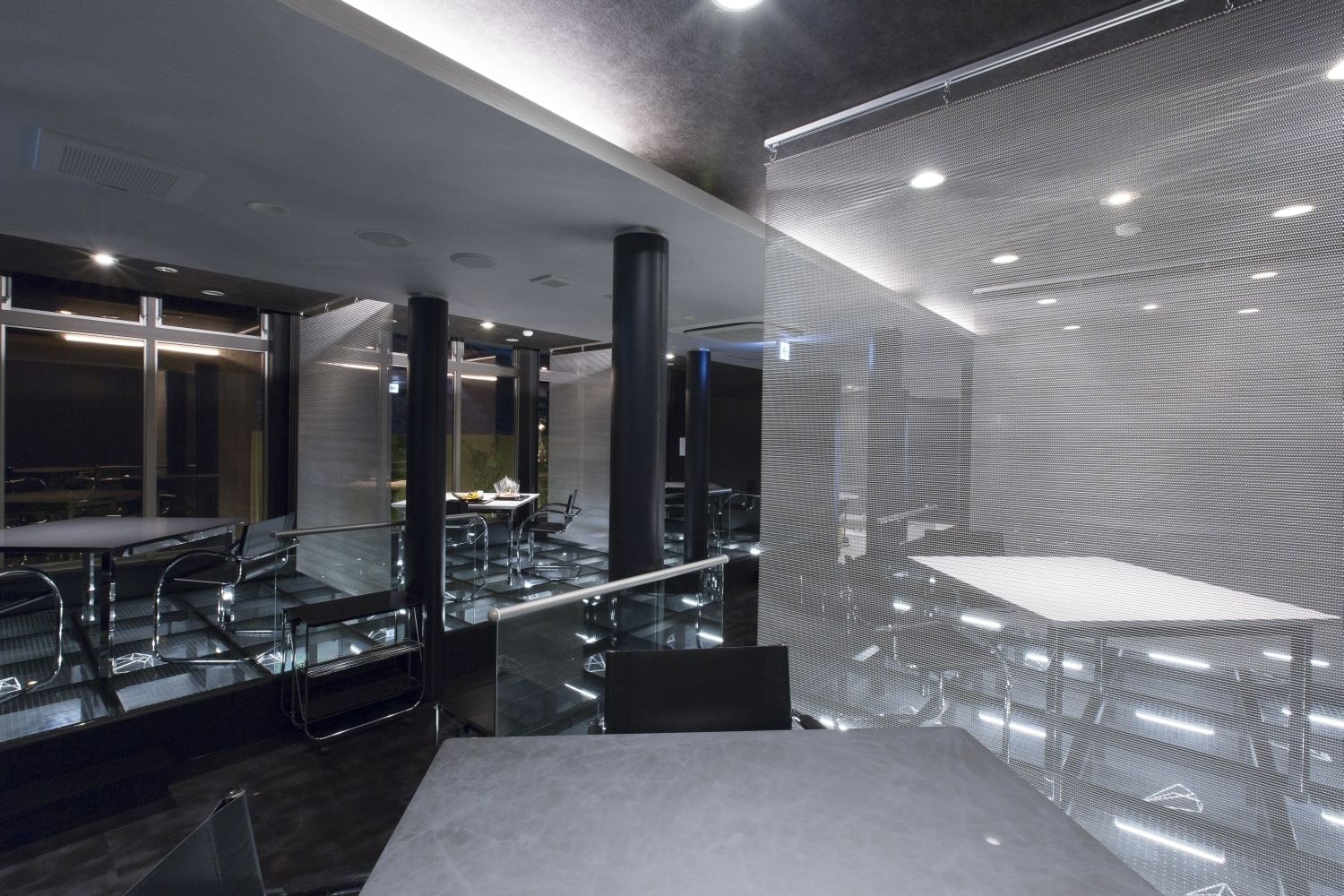 A modern hotel lobby with a glass staircase, a seating area with tables and chairs, and a large wall with a geometric pattern.