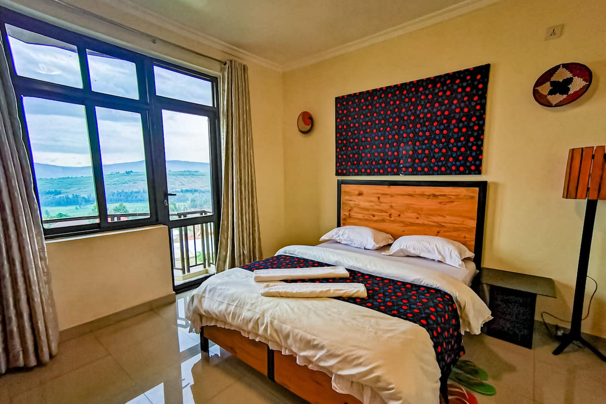 A hotel room with a large window, a bed with white linens, a wooden headboard, and a view of the countryside.