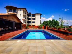 A modern pool area with a clear blue pool, wooden deck, and a view of a multi-story building with balconies. - Peponi Livingspaces (Kigali)