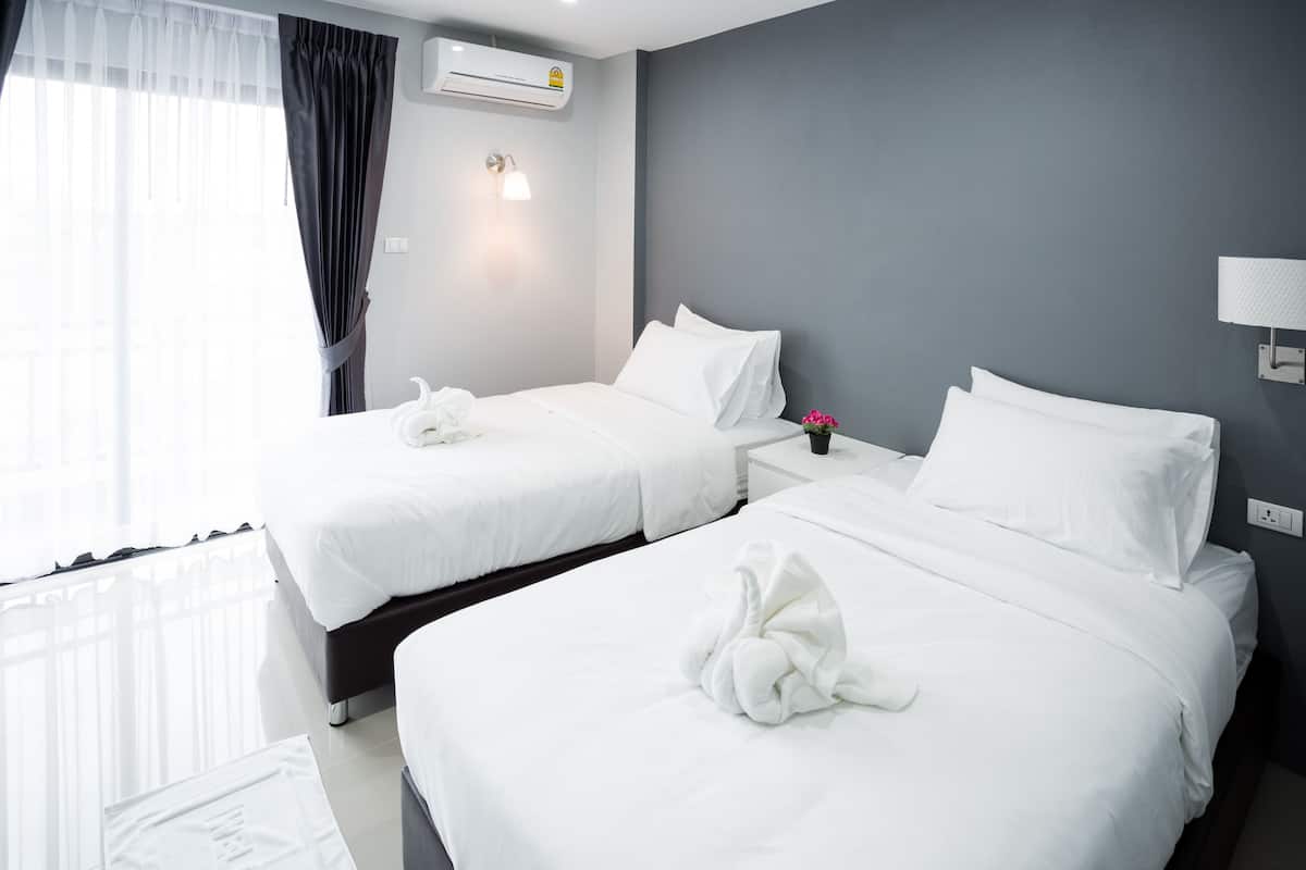 Quarto de hotel com duas camas, roupa de cama branca, mesa de cabeceira com abajur e ar condicionado na parede.