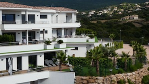 A multi-story building with balconies, outdoor seating, and a view of a hilly landscape.