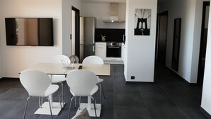 A modern kitchen with stainless steel appliances, a dining area with white chairs and a table, and a large flat-screen TV mounted on the wall.