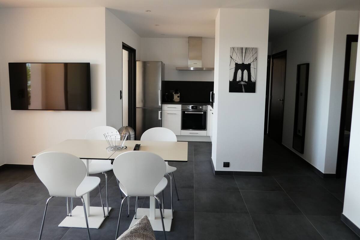 Een moderne keuken met roestvrijstalen apparatuur, een eethoek met witte stoelen en een tafel, en een grote flatscreen televisie aan de muur bevestigd.