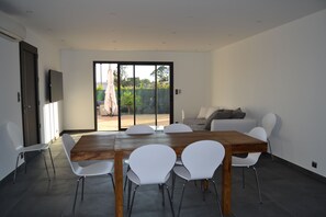 Uma sala de jantar moderna com uma mesa de madeira e cadeiras brancas, um sofá e uma TV fixada na parede.