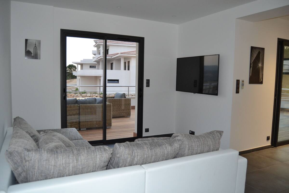 Een moderne woonkamer met een witte bank, grijze kussens en een grote schuifdeur naar een buitenbalkon met rieten meubels.