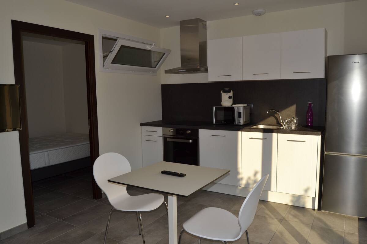 Een kleine keuken met een magnetron, oven en koelkast. Er is een eettafel met twee stoelen.