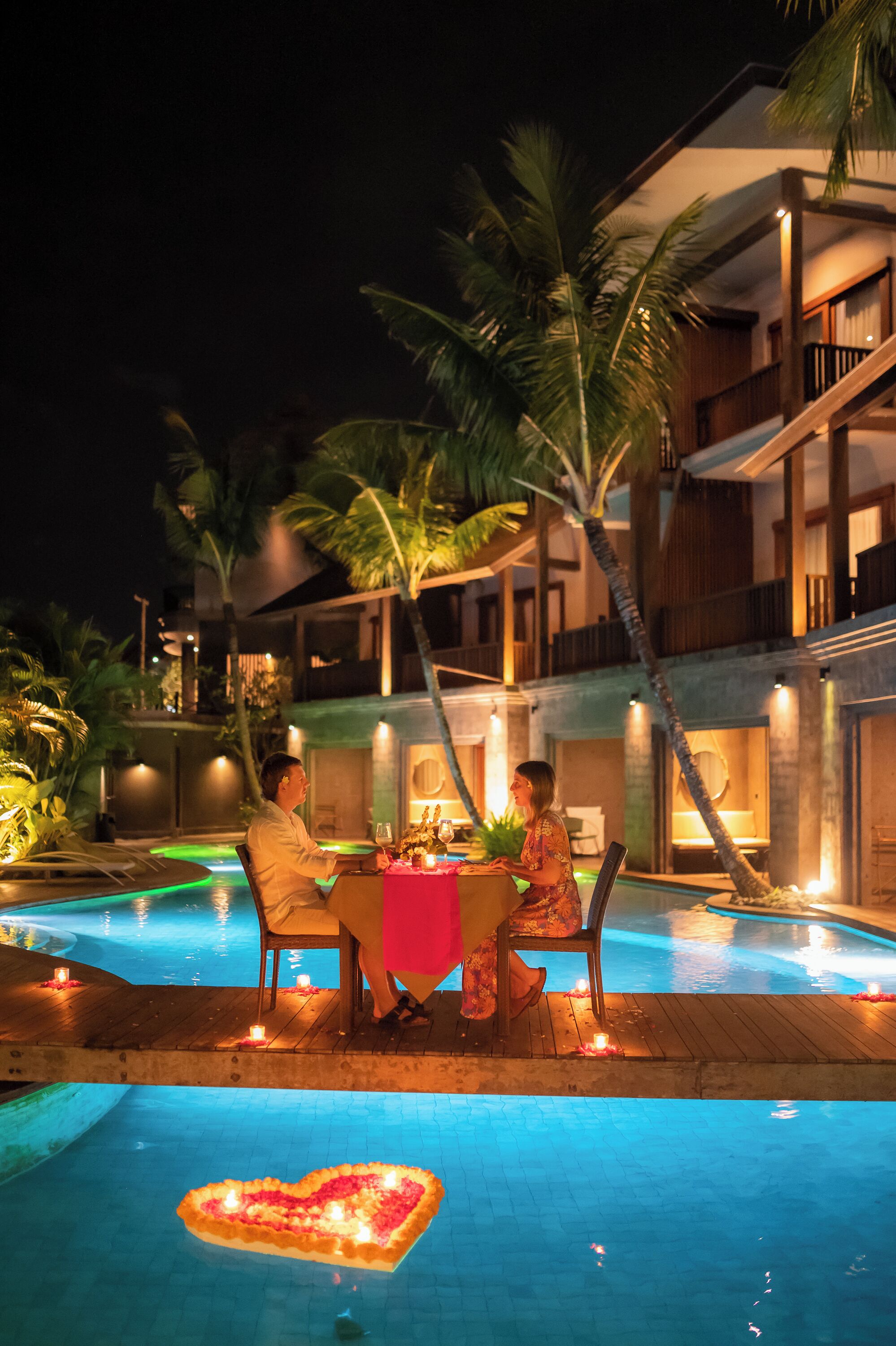 A couple dining at a poolside table with a heart-shaped arrangement of flowers on the water.