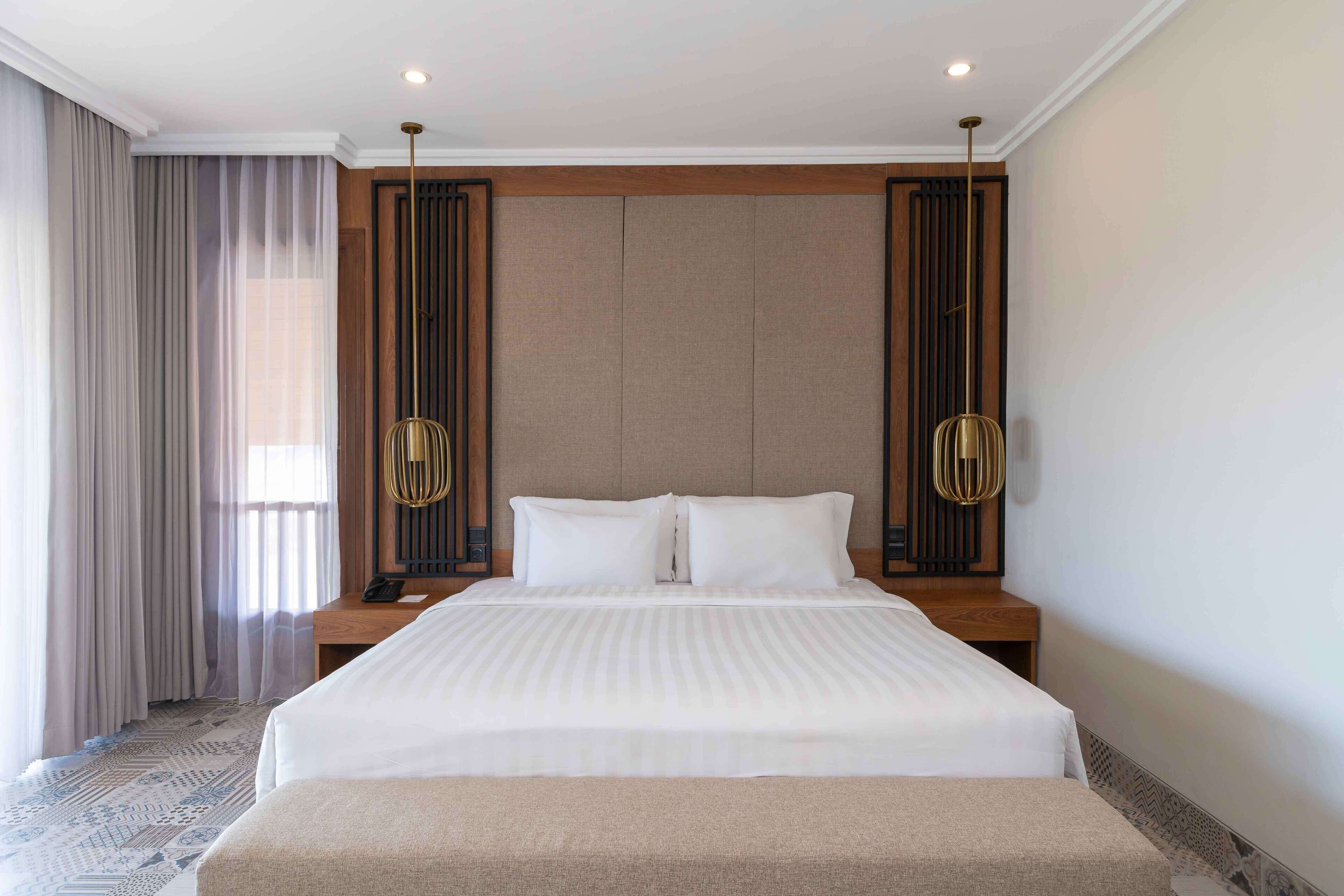 Habitación de hotel con una cama grande, cabecera de madera, mesita de noche y un amplio ventanal con cortinas.