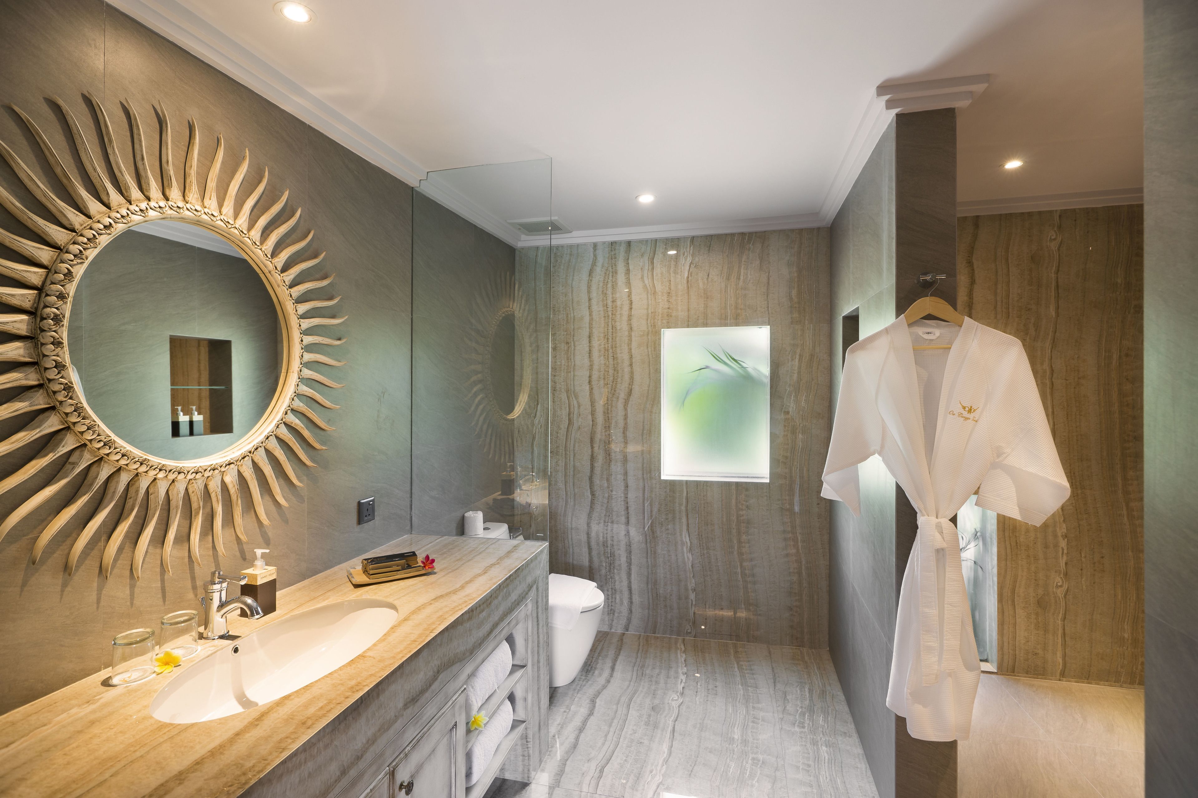 A modern bathroom with a sunburst mirror, a wooden vanity, a white toilet, and a robe hanging on the wall.