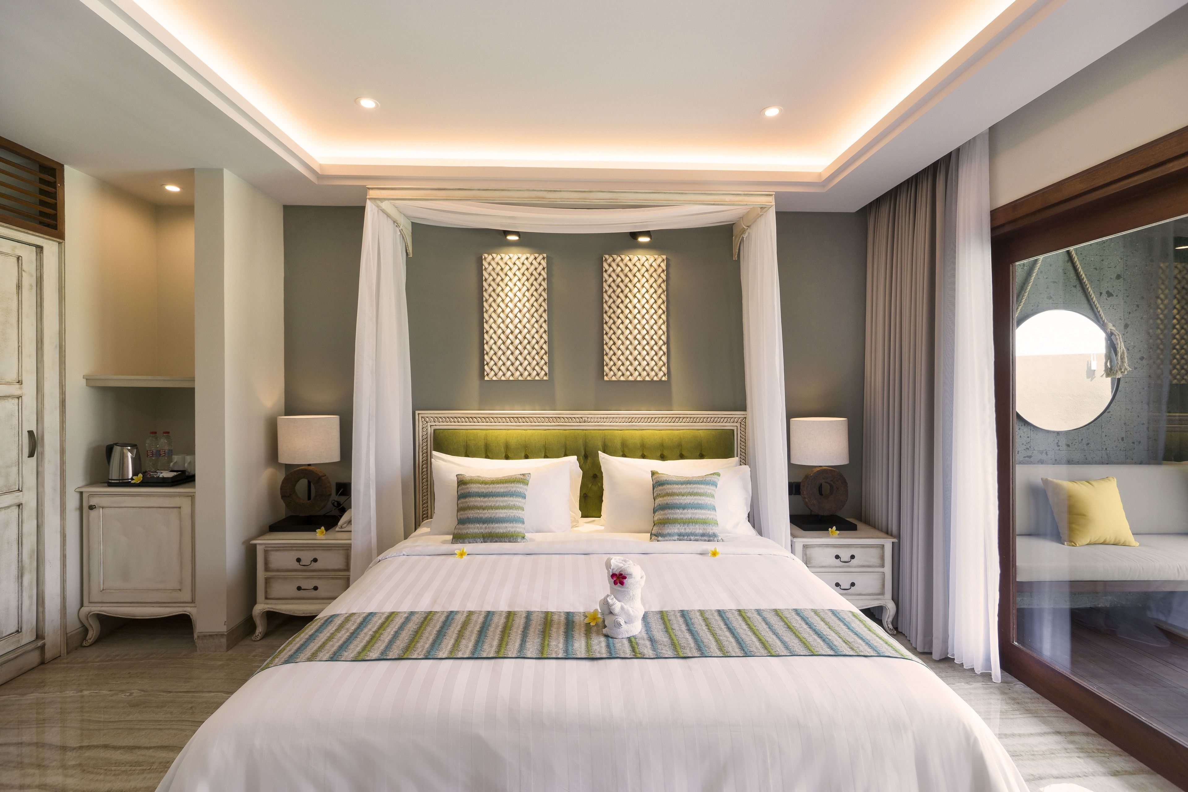 A large bed with a canopy, two bedside tables with lamps, a nightstand, a decorative rug, and a view through a glass door.