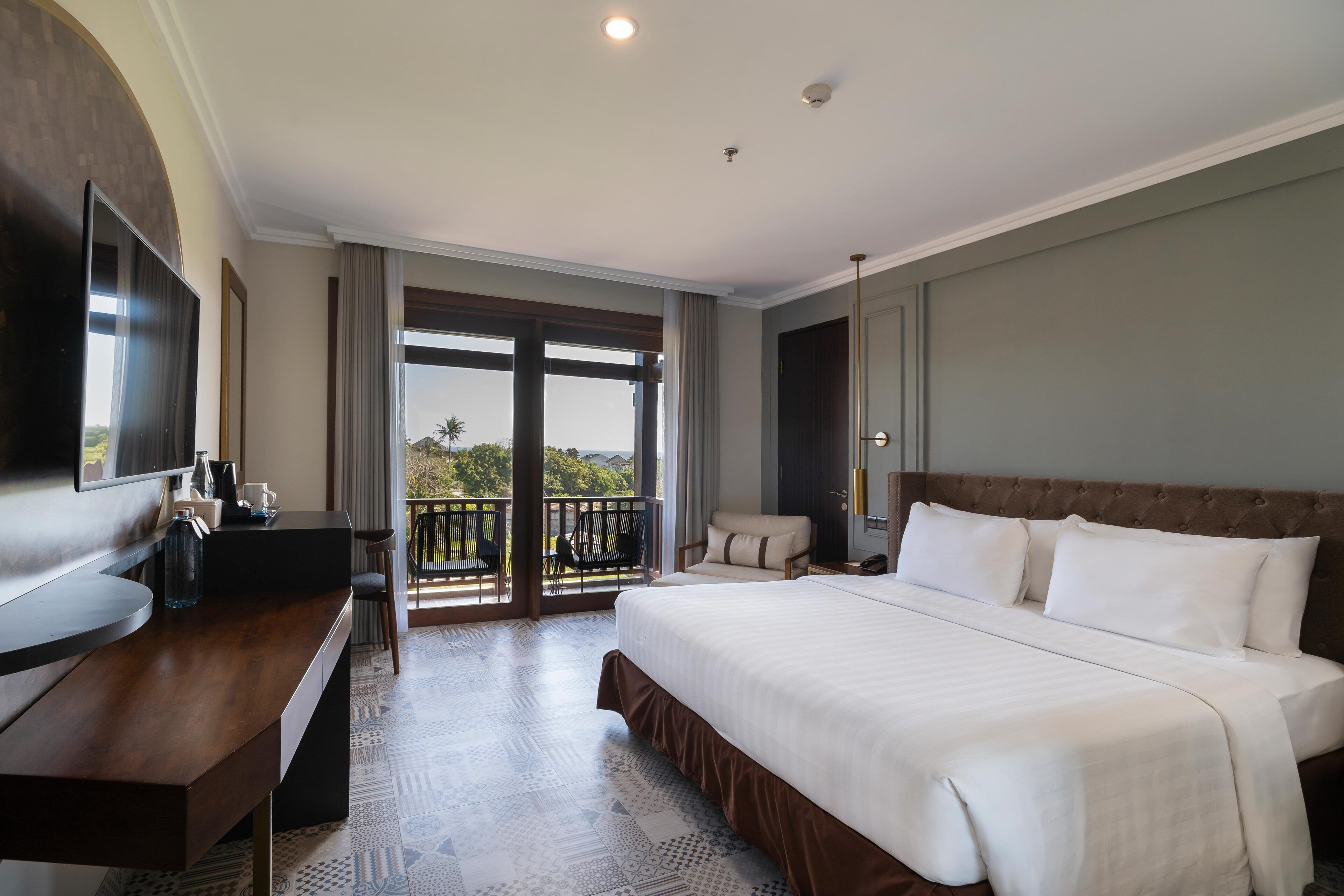 A hotel room with a large bed, a desk with a flat-screen TV, a balcony with a view, and a large window.