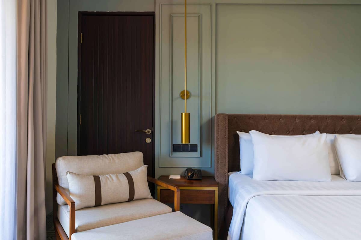 A hotel room with a bed, a chair, a bedside table with a phone, and a door.