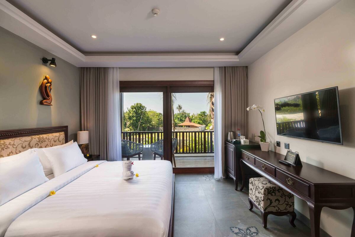 A hotel room with a large bed, a desk, a television, and a balcony with a view of greenery.