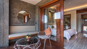 Una habitación moderna con pared de piedra, un espejo redondo, una silla blanca, una mesa de vidrio con frutero y una cama con ropa de cama blanca.