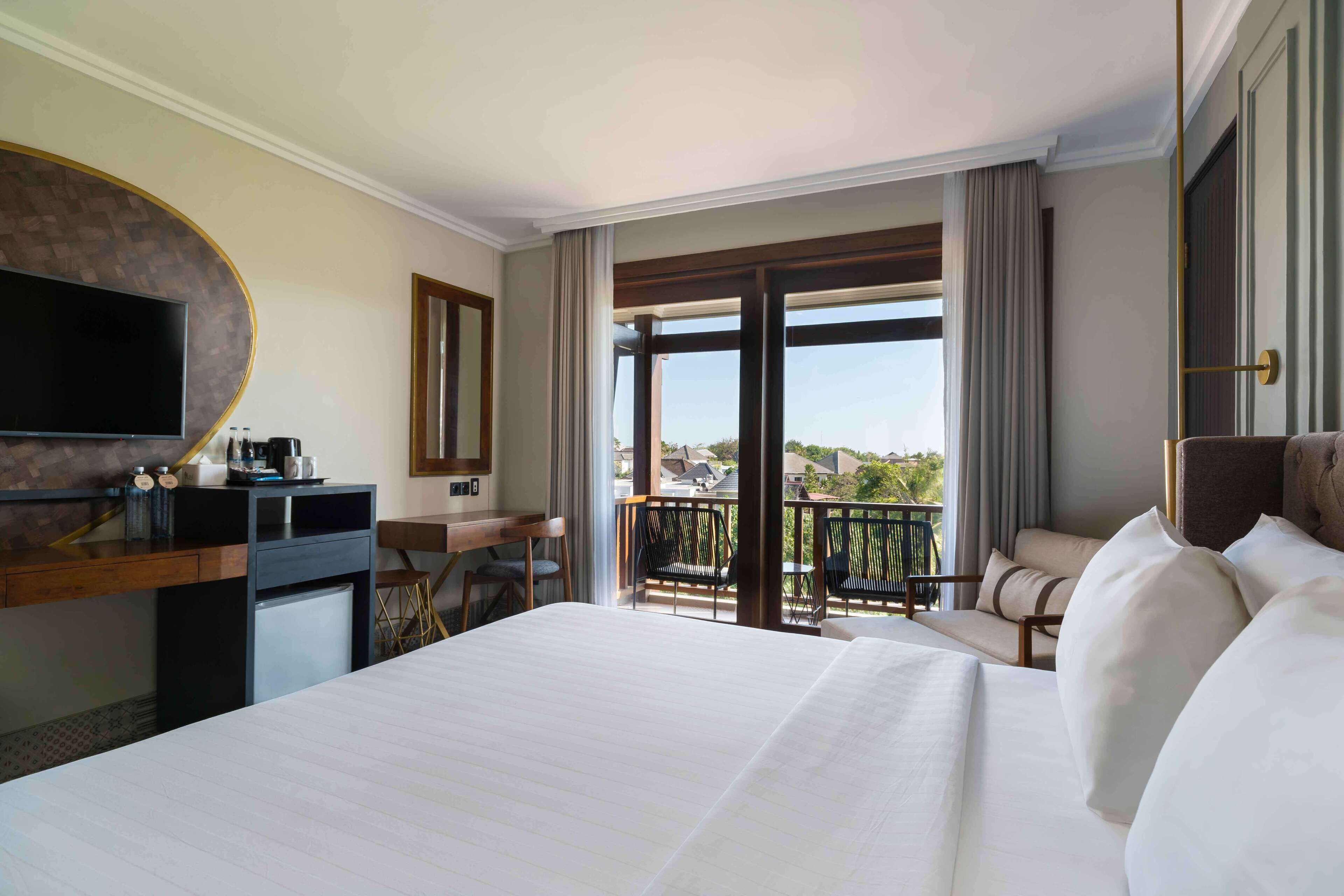 A hotel room with a large bed, a desk with a flat-screen TV, a small fridge, and a balcony with a view of the city.