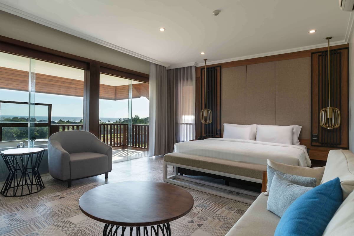A spacious hotel room with a large bed, a sofa, a round table, and a balcony with a view.