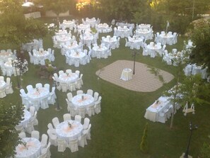 Un salón de bodas al aire libre con varias mesas redondas dispuestas en círculo, rodeadas de sillas blancas.