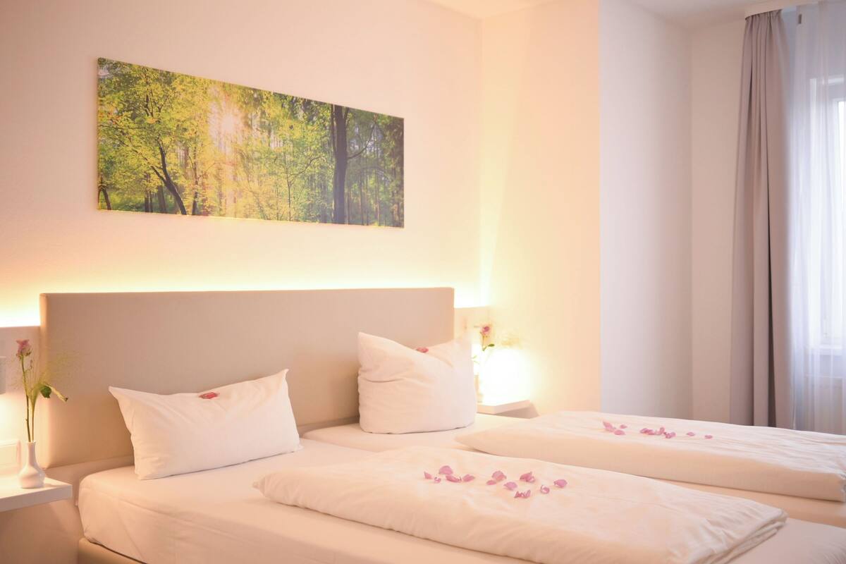 Habitación de hotel con dos camas, un cuadro grande de un bosque y una mesita de noche con un jarrón de flores.