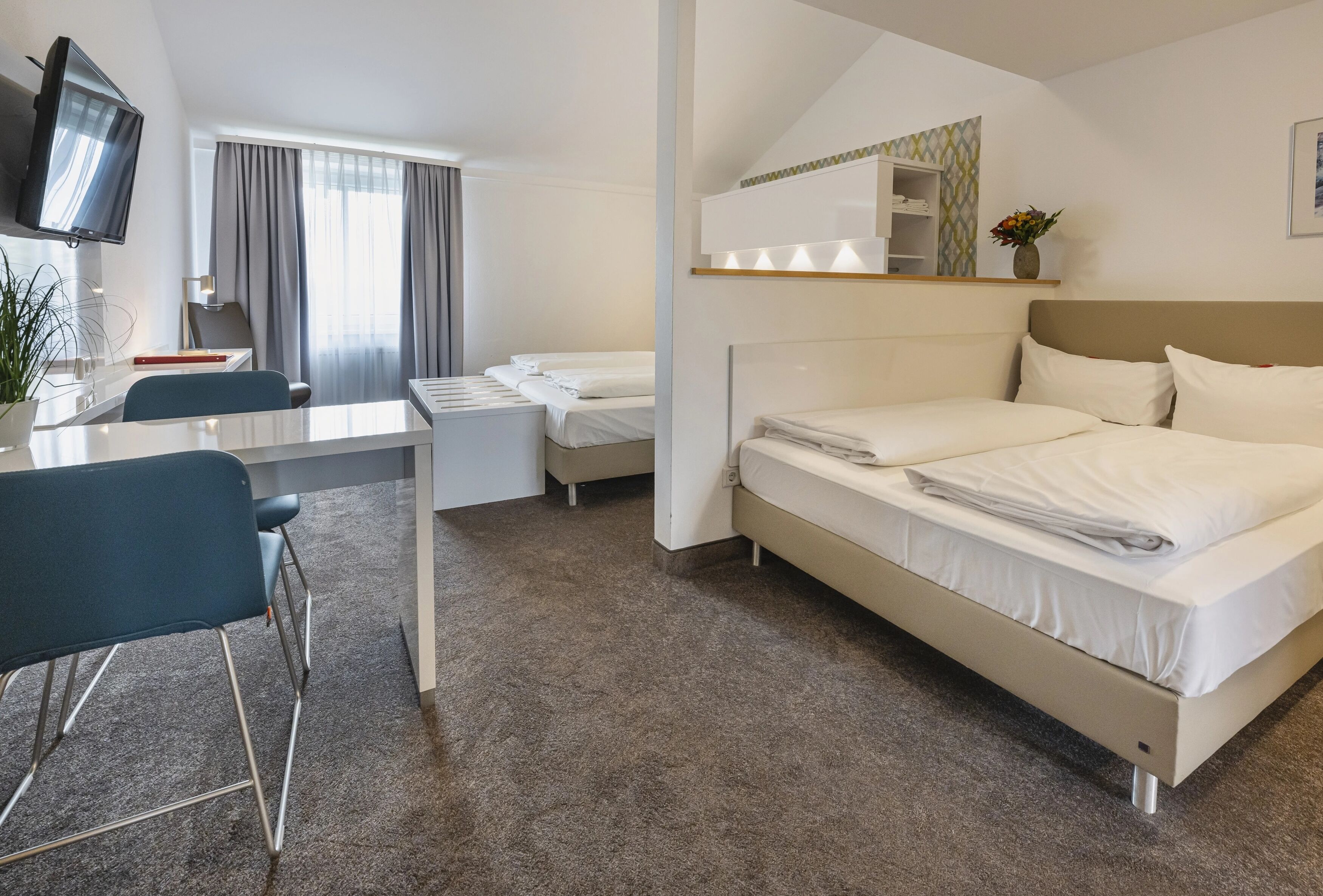 A hotel room with a bed, a desk, a chair, a TV, and a small shelf with a vase.