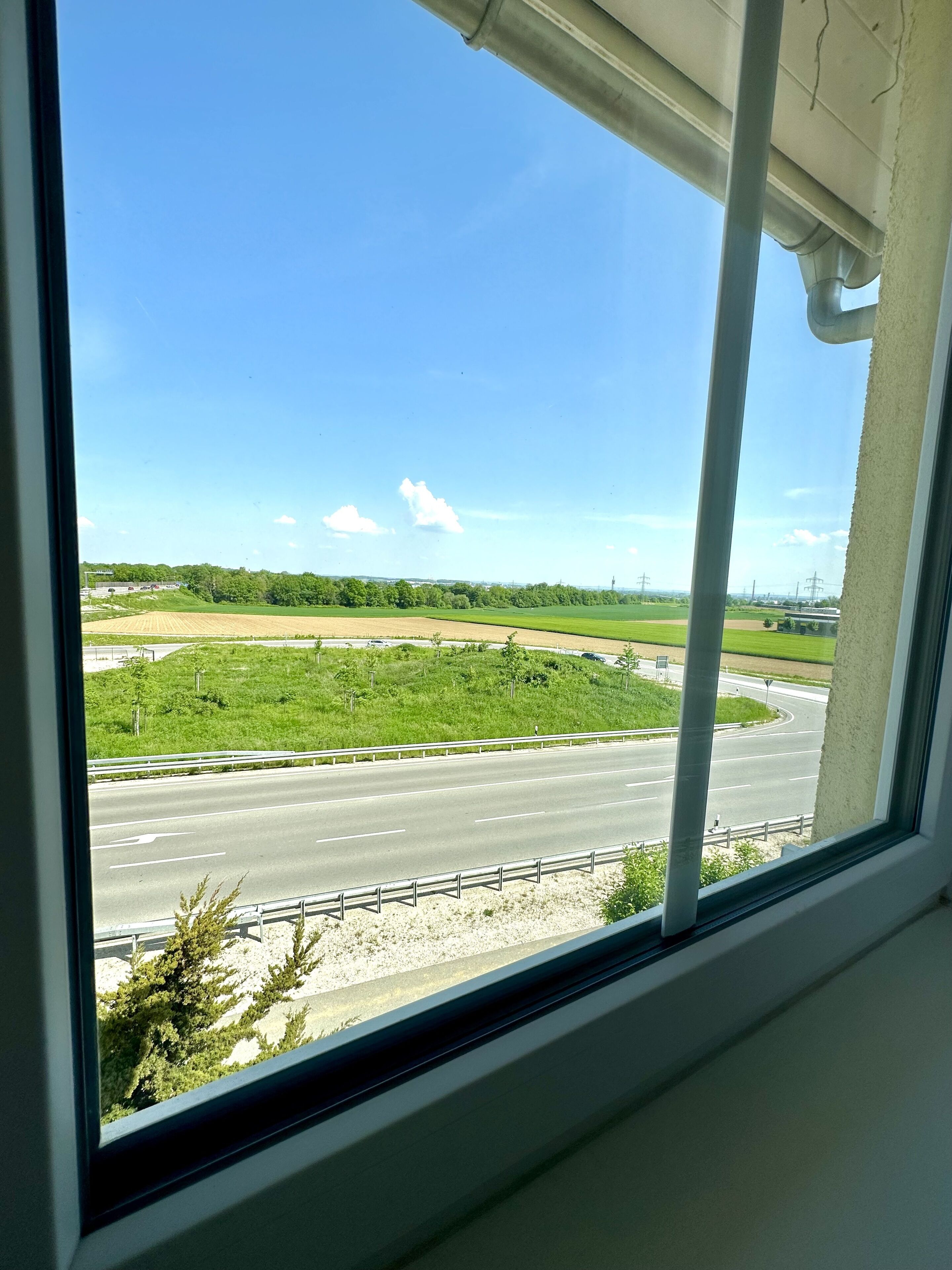 Vista desde una ventana sobre una autopista, con un área verde y árboles a lo lejos.