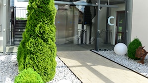 Una entrada moderna a un edificio con puertas de cristal, un seto alto de color verde y una escultura blanca esférica.