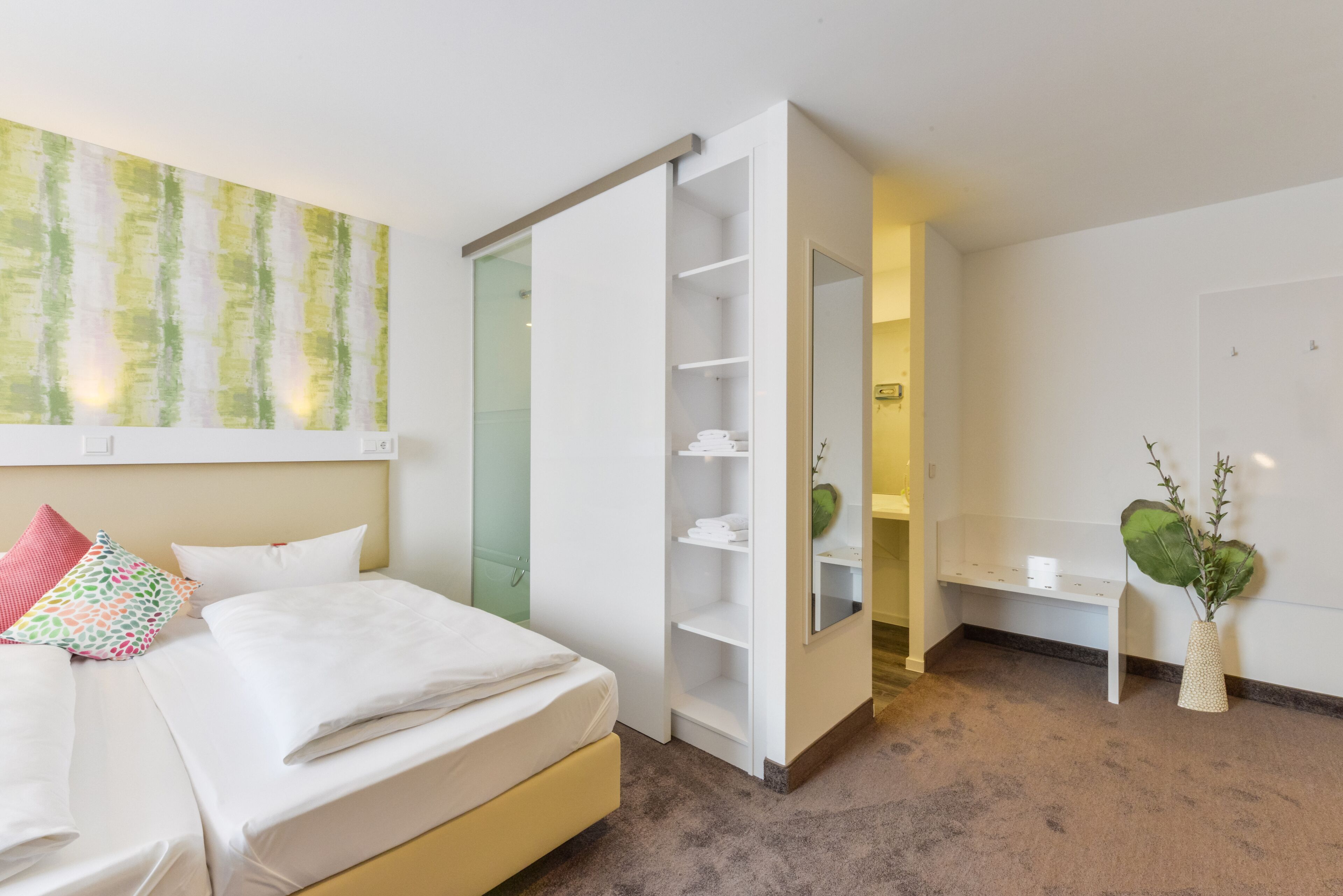 Una camera d'albergo con un letto, un armadio bianco, una panca e una pianta verde.