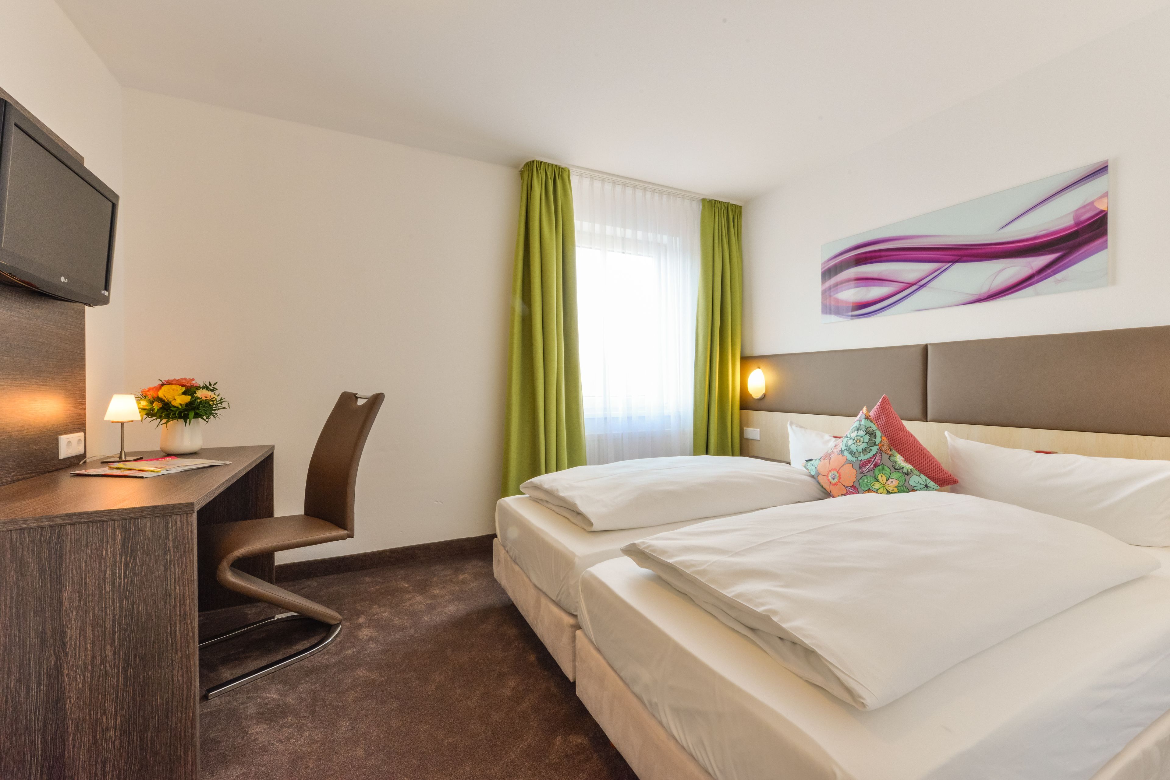 A hotel room with a desk, a chair, a bed with pillows, a TV, and a window with curtains.