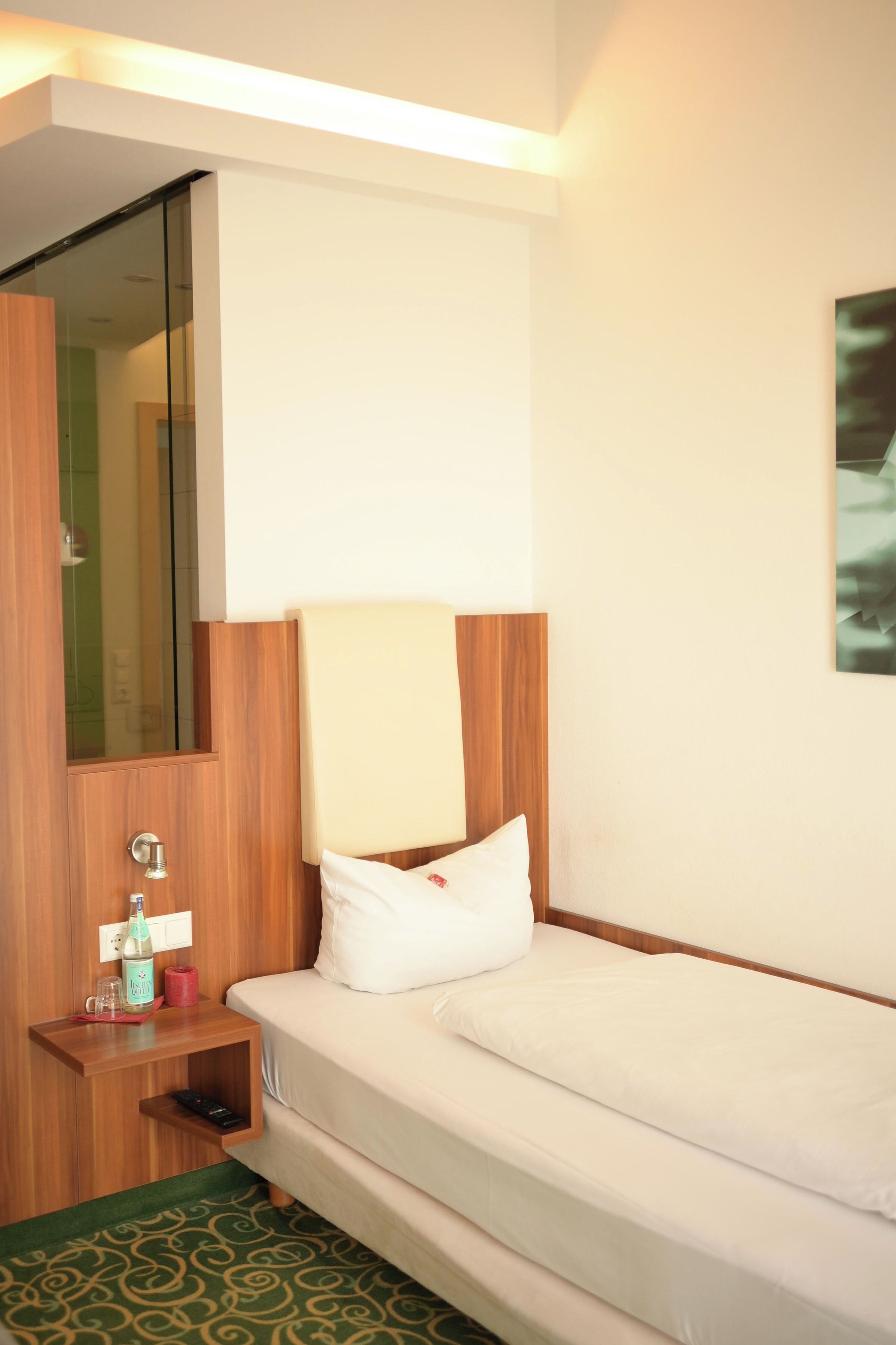 A hotel room with a bed, a nightstand, a mirror, and a green patterned rug.