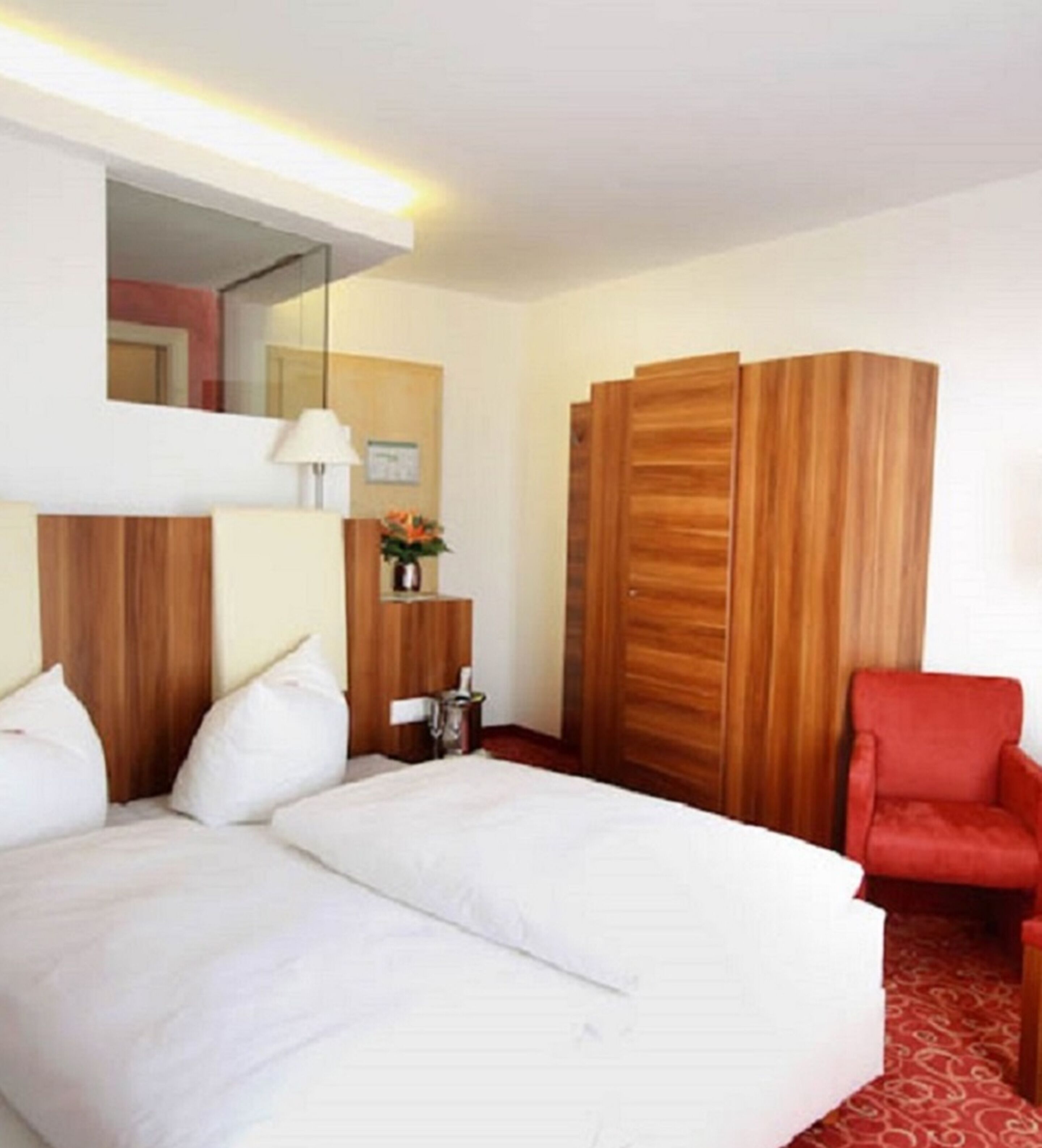 A hotel room with a bed, a red chair, a wooden wardrobe, a mirror, and a small table with a vase.