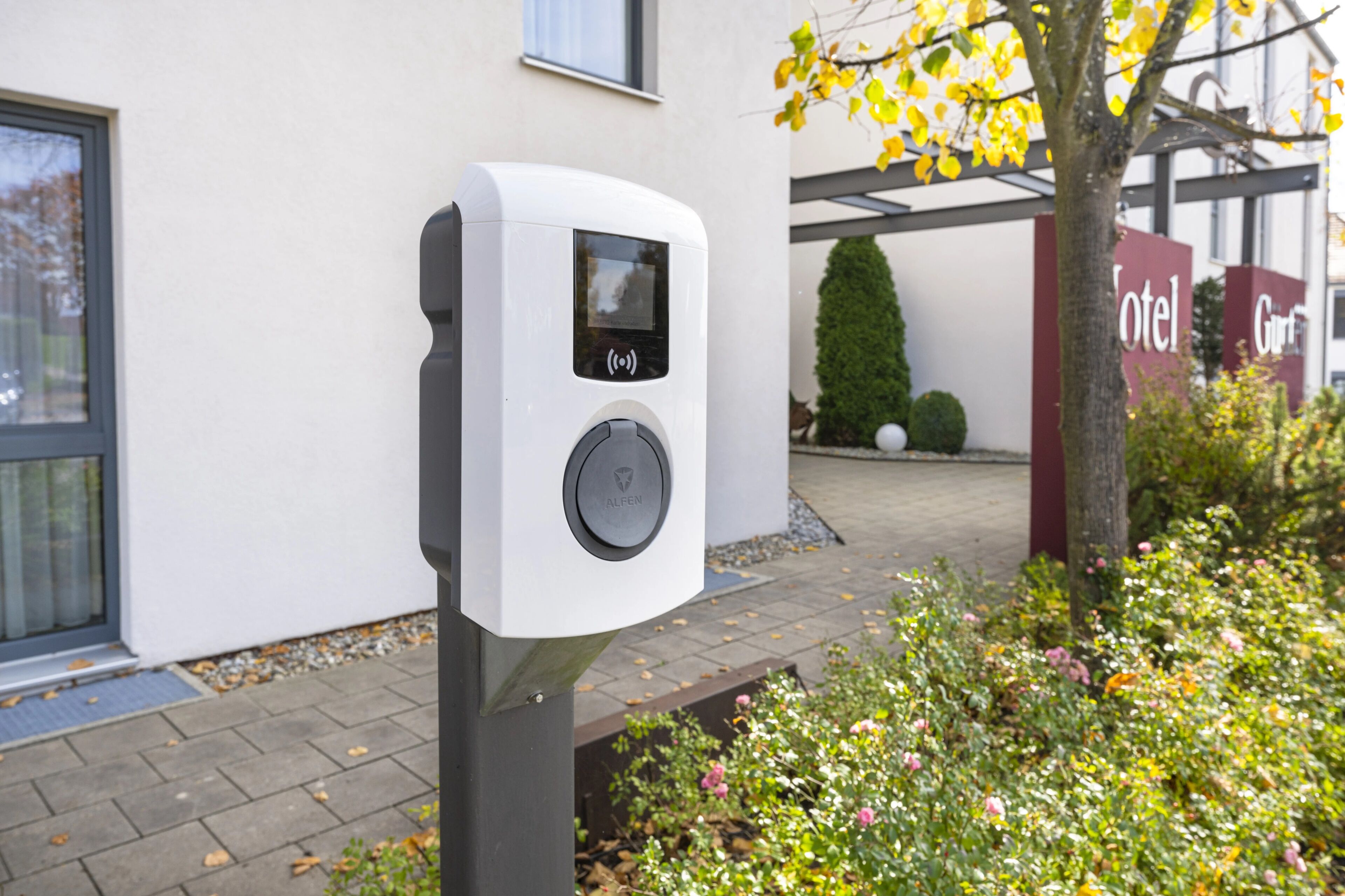 A white electric vehicle charging station with a screen and a button, located outside a building with a sign that reads 'Hotel.'