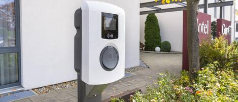 A white electric vehicle charging station with a screen and a button, located outside a building with a sign that reads 'Hotel.'