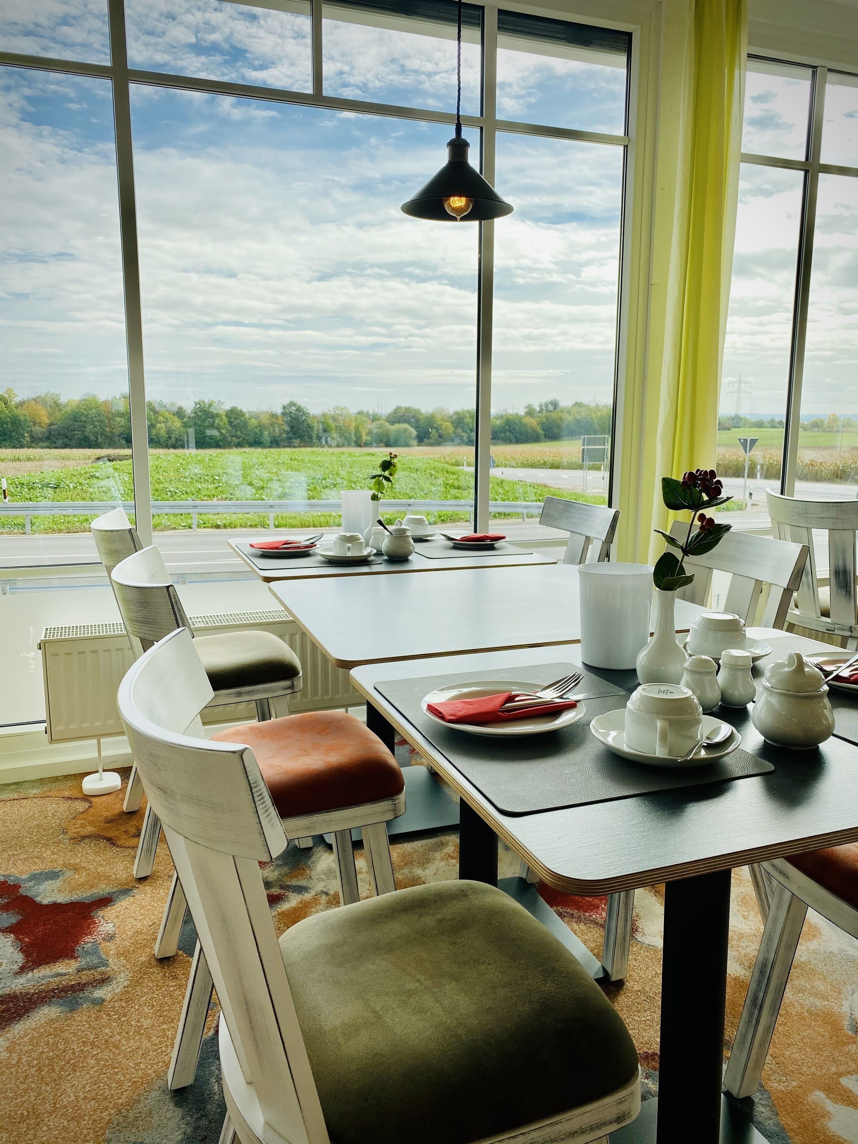 Ein Essbereich mit Tisch für zwei, Stühlen, einer Hängelampe und Blick auf ein grünes Feld durch große Fenster.