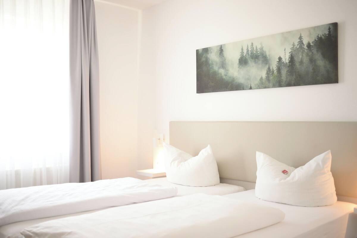 Habitación de hotel con cama, ropa de cama blanca, cabecera, mesita de noche, lámpara y un cuadro de un bosque en la pared.