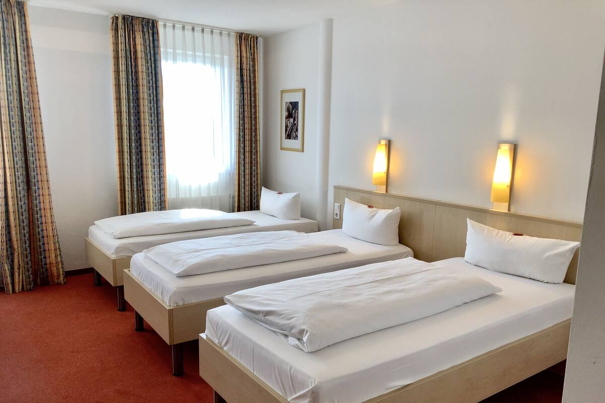 Habitación de hotel con tres camas individuales, alfombra roja, ventana con cortinas y dos luces fijadas a la pared.
