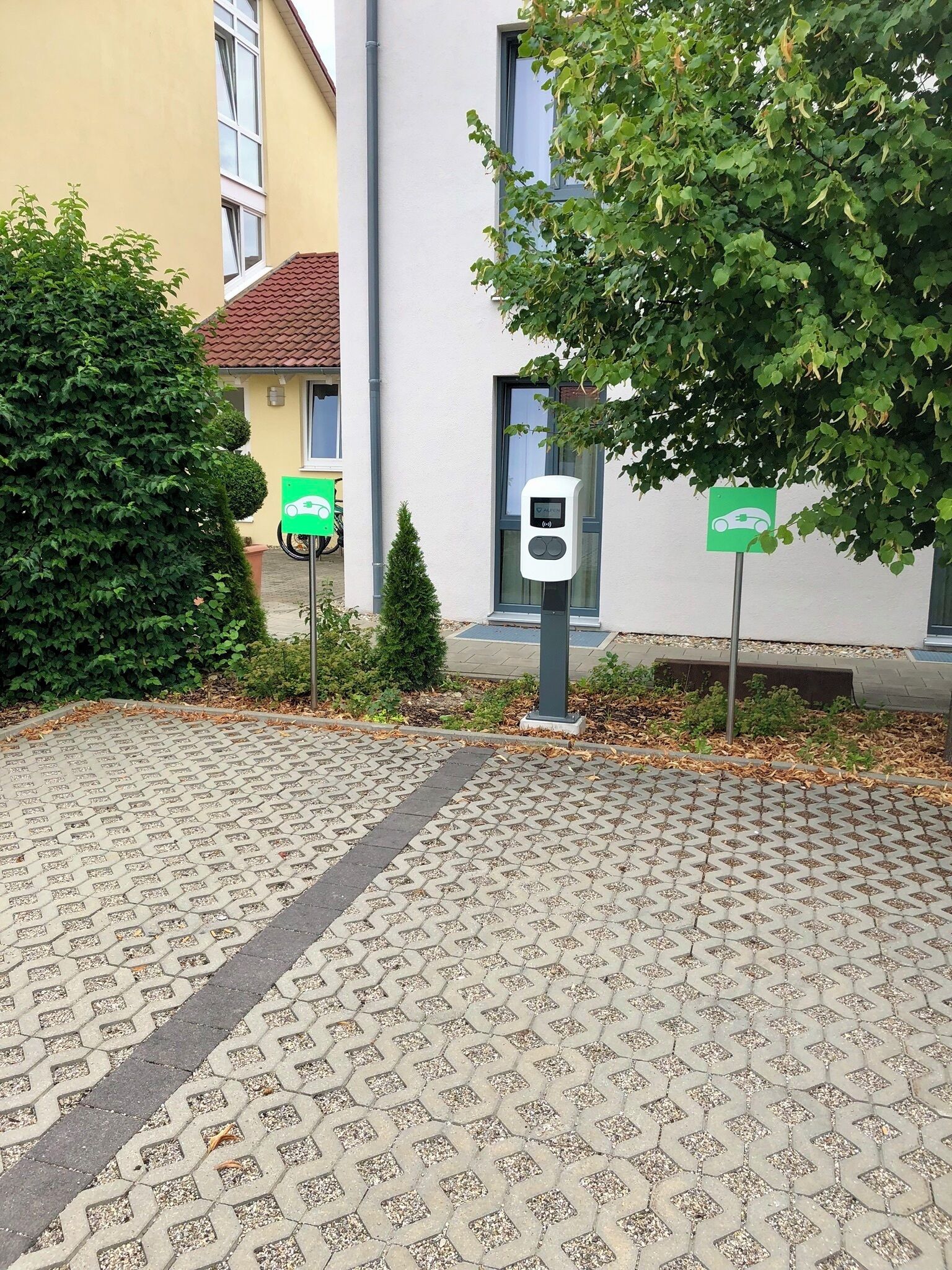 A parking area with electric vehicle charging stations, a paved walkway, and a building with a tiled roof.