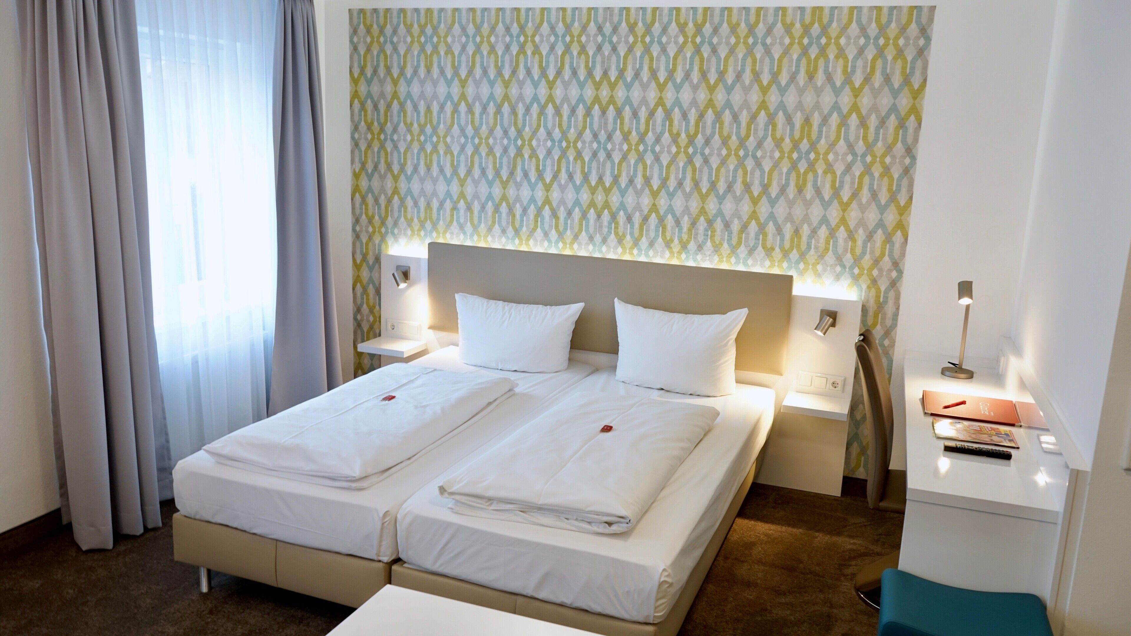 A hotel room with a double bed, two bedside tables, a desk with a chair, and a patterned wall.