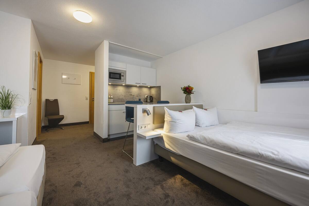 Una habitación de hotel con cama, escritorio, silla, kitchenette y televisión.