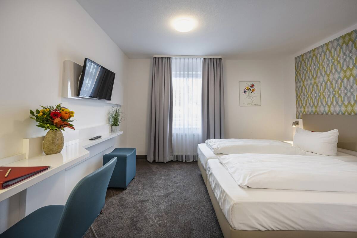 Habitación de hotel con cama, escritorio, silla, televisor y ventana con cortinas.