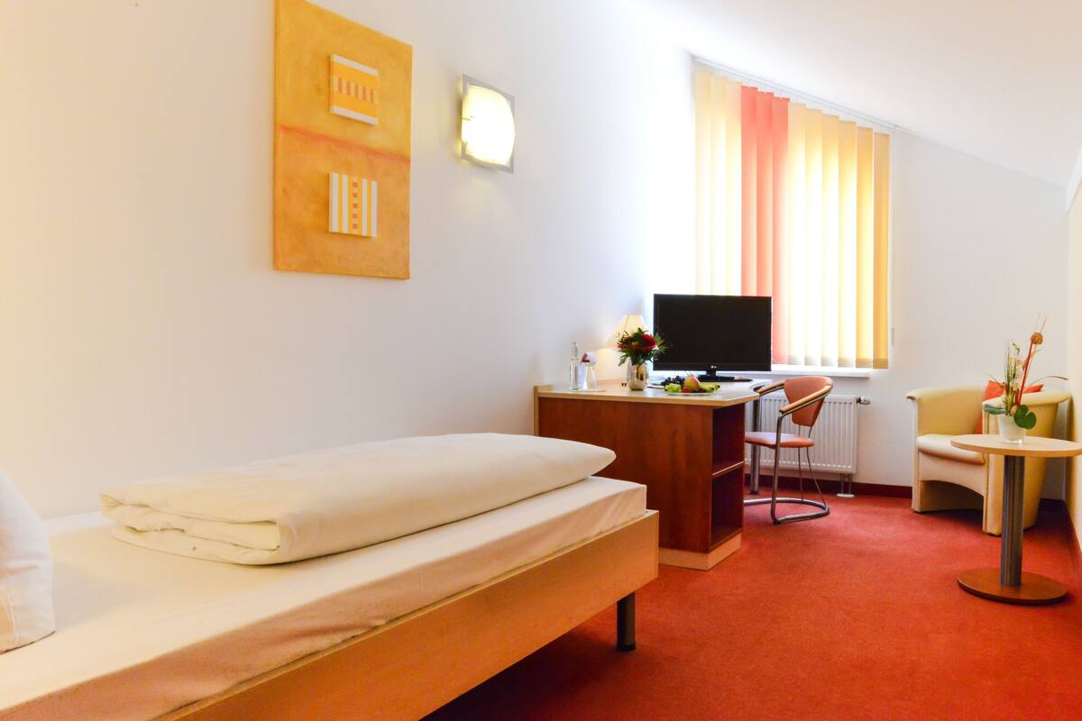 Habitación de hotel con cama, escritorio con computadora, silla, mesita y ventana con cortinas.