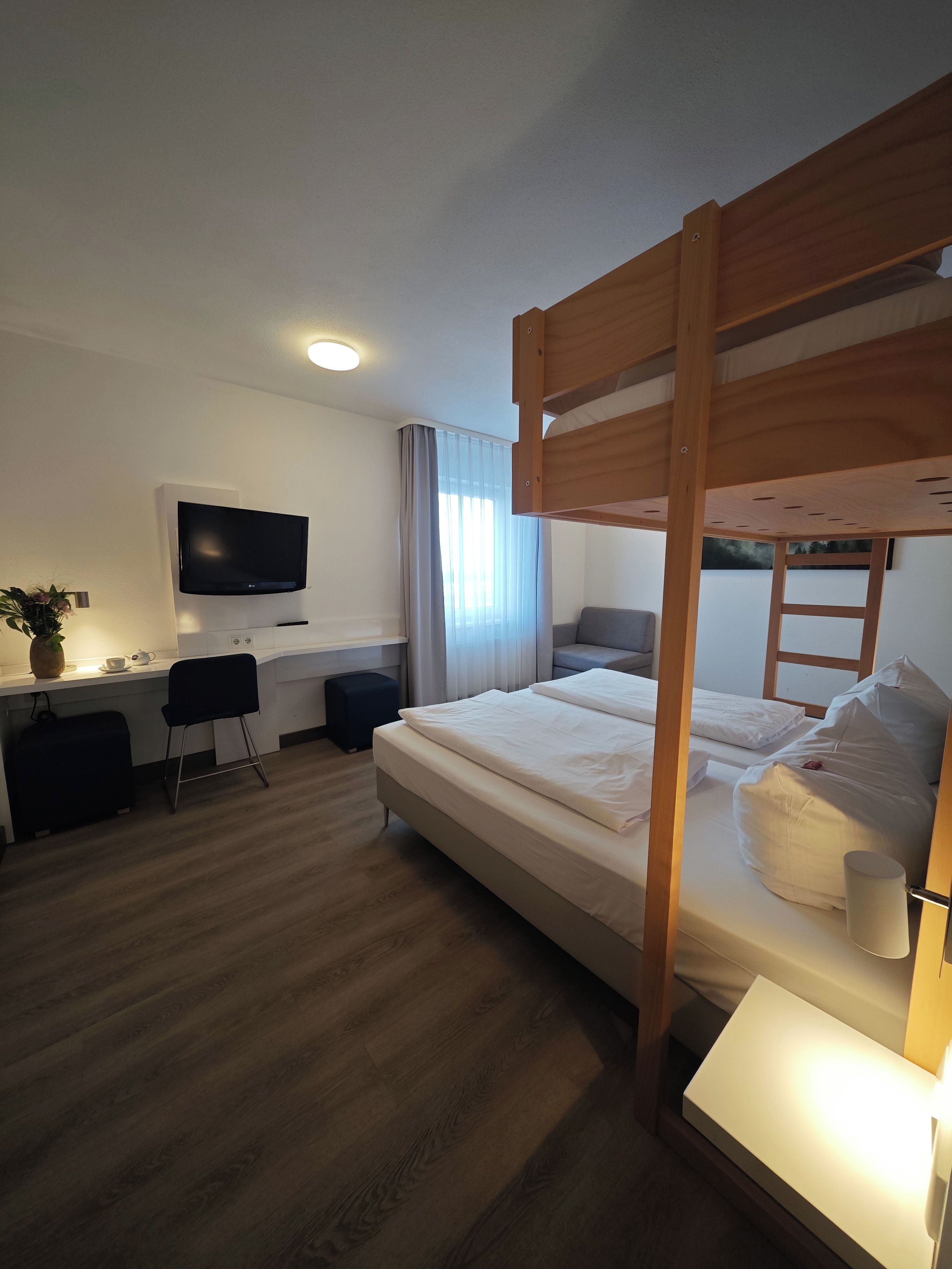 A bunk bed with a wooden frame, a desk with a chair, a television, and a window with curtains.